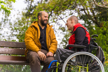 Portrait of a paralyzed young man in a wheelchair with a friend on a bench in a public park in the city, talking and laughing