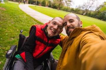 Selfie of a paralyzed young man in a wheelchair with a friend on a bench in a public park in the city, talking and laughing