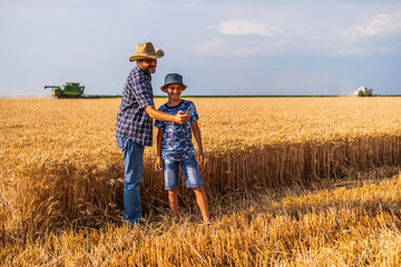 Naklejka na ściany i meble Farmers are standing in their wheat field while the harvesting is taking place. Father is teaching his son about agriculture.