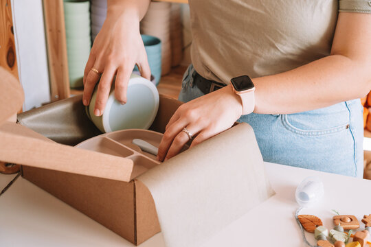 Cropped image of woman hands packing and wrapping shipment with silicone kids dishes in cardboard box for delivery. Wooden rattle toy, trinket decoration nearby on desk. Workshop store, work at home