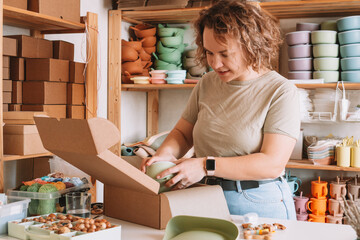 Smiling concentrated curly woman packing and wrapping shipment with silicone baby dish in cardboard...