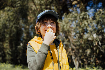 Smiling cheerful child kid in yellow vest and green hoodie eats crisp snacks outdoors in public park. Schoolboy boy enjoying consumes chews junk food outside with trees vegetation on the background.