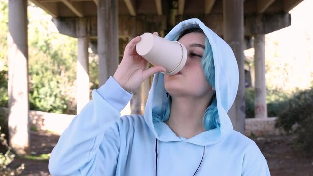 Teenage girl in blue oversize hoodie drinking coffee to go. Blue haired teen girl staying outdoors against bridge pillars. Mockup, head shot. Hipster and adolescence concept
