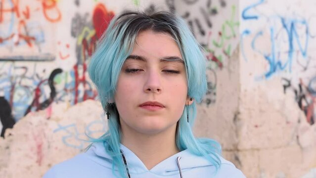 Serious Teenager in light blue oversize hoodie. Blue haired teen girl staying outdoor against graffiti wall and looking into camera. Head shot
