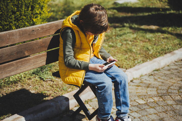 Schoolboy plays game his cell phone sitting on bench in the city park. Boy kid in yellow vest and green hoodie uses mobile gadget to chat with friends while outdoors in the garden during sunny day.
