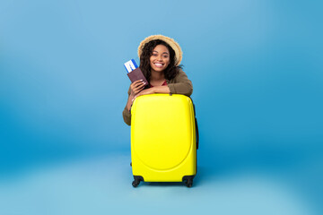 Happy black lady in casual wear sitting behind suitcase, holding passport and flight tickets, going on summer vacation
