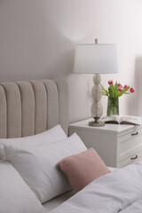 Stylish lamp, flowers and magazine on bedside table indoors. Bedroom interior elements