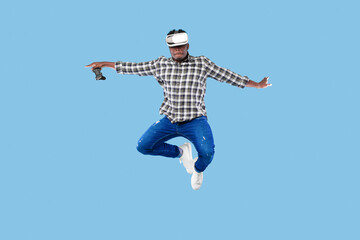 Fototapeta na wymiar Young African American guy in VR headset holding joystick, jumping up in air on blue background
