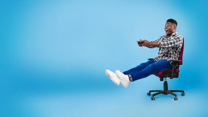 Excited young black guy with joystick sitting on chair, playing video game on blue background, banner with copy space