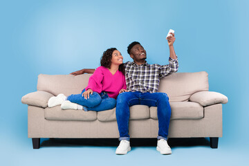 Split system. Young black couple with remote relaxing under air conditioner, sitting on couch over...