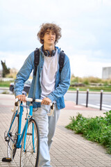 Young curly-haired student with headphones walking to college holding bicycle with hands.