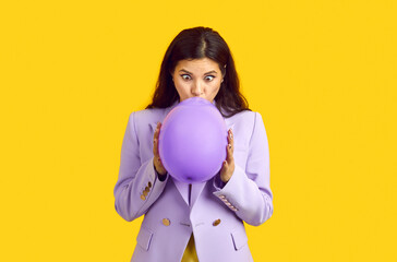 Beautiful young woman in suit blowing up balloon. Long haired brunette lady in lilac jacket standing isolated on bright yellow background and inflating purple balloon with funny face expression