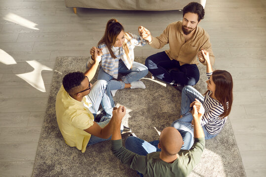 Diverse group of people having bonding time. High angle indoor shot of young mixed race friends sitting in circle on floor, holding hands and meditating together. Community and friendship concept - Powered by Adobe