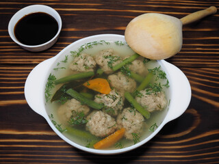 Hot meatball soup with green beans, carrots and herbs in a white bowl on a wooden table, soy sauce, spoon, top view, flat layout. Transparent broth with meat and vegetables for dietary nutrition