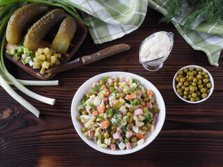 Mixed vegetable salad olivier and canned peas in bowls, salted cucumbers, green onions, mayonnaise sauce on a dark wooden table, flat layout. Traditional salad recipe for lunch, Russian cuisine