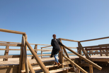 A red-haired woman in a long black jacket, climbing some steps of a wooden walkway, on the beach of Isla Cristina, Spain.