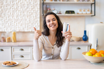 Glad woman holding glass with clean fresh water and showing thumb up, sitting in kitchen interior and smiling at camera