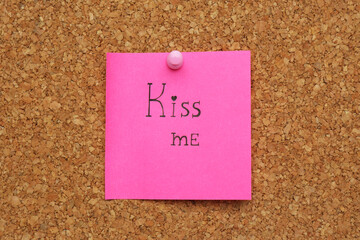 Pink paper note with phrase Kiss Me pinned to cork board