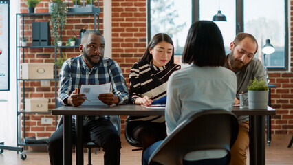 Multiethnic group of HR workers interviewing female candidate about job offer, analyzing resume information on papers. People doing teamwork at job interview with recruitment applicant.