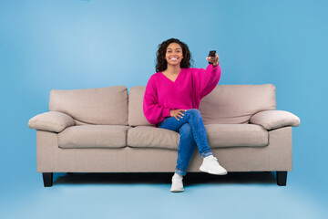 Obraz premium Young African American lady with remote control sitting on couch and watching TV on blue studio background, copy space