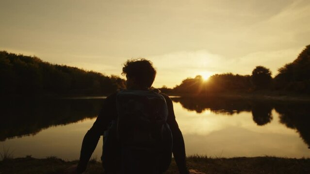 boy with backpack sits near the lake at sunset