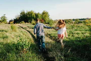 Fototapeta premium Rear view of a romantic man and woman walking on field grass, nature enjoying sunlight. Concept of lovely family holding hands. Young couple running and looking away. Back view.