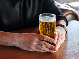 Hands of a man holding a glass of beer on the bar or pub table.