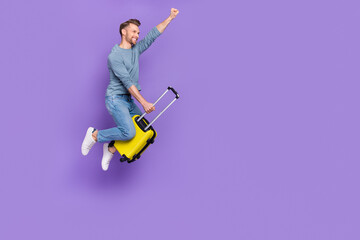 Photo of excited cool guy dressed grey shirt jumping high riding luggage fist empty space isolated purple color background
