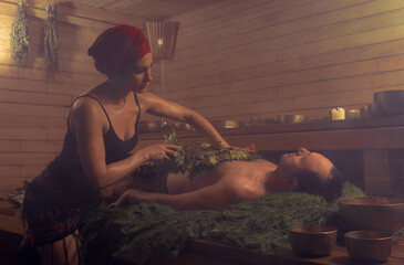 A woman conducts a bathing ritual for a man with the help of brooms. A man lies on spruce branches