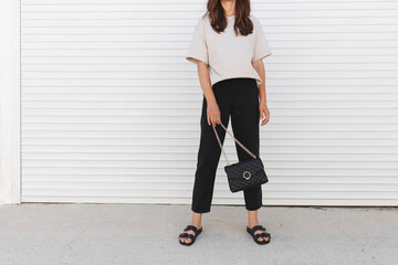 Woman wearing beige t-shirt, black pants, bag with chain and flat sandals walking outdoor near white roller door. Details of stylish trendy basic minimalistic casual outfit. Street fashion. No face.