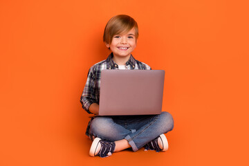 Full size photo of young boy sit floor use laptop study online isolated over orange color background