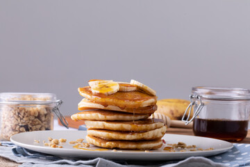 Pancakes with banana slices and syrup served in plate by oats and honey jar against gray background