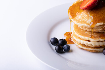 Close-up of dripping syrup over stacked pancakes with berry fruits in plate on white background