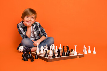 Full length photo of young boy hand touch chin look empty space hobby pay game sit floor isolated over orange color background