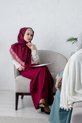 Portrait of a Muslim woman psychologist dressed in a burgundy hijab holds a tablet in her hands and...
