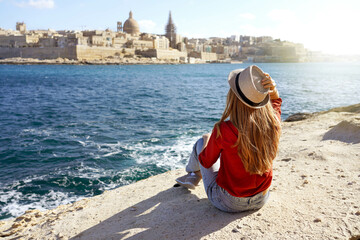 Beautiful young woman with hat sitting on stone by the sea looking at stunning panoramic view of Valletta city in Malta - obrazy, fototapety, plakaty