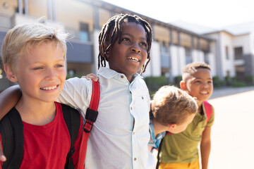 Smiling multiracial elementary schoolboys with arm around standing at school campus during sunny day