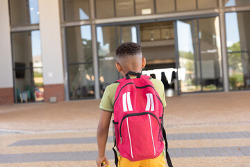 Rear view of biracial elementary schoolboy with backpack standing against school building