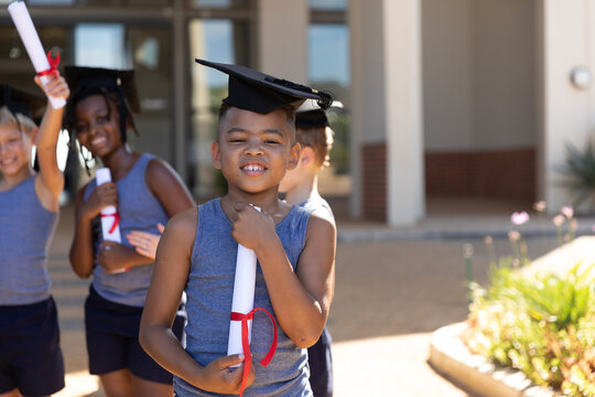 Biracial elementary schoolboy wearing mortarboard while holding degree with classmates in background