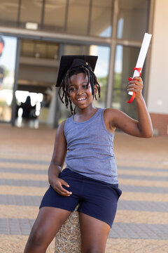 Happy african american elementary schoolboy with mortarboard holding degree while sitting on pole