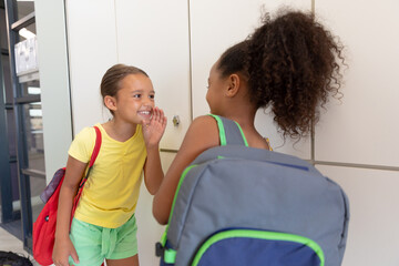 Multiracial elementary schoolgirls gossiping while standing by lockers in school corridor