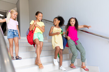 Full length of multiracial elementary schoolgirls moving down steps in school building - obrazy, fototapety, plakaty