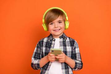 Photo of young cheerful boy listen music earphones melody app mobile isolated over orange color...
