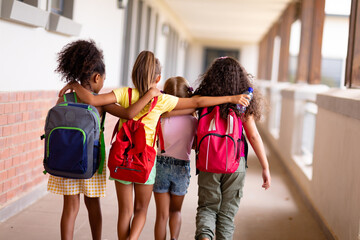 Fototapeta Rear view of multiracial elementary schoolgirls with backpacks and arm around walking in corridor obraz