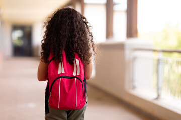 Rear view of biracial elementary schoolgirl with backpack walking in corridor at school - Powered by Adobe
