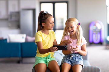 Caucasian elementary schoolgirl showing smart phone to caucasian friend sitting on couch