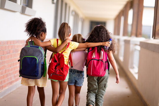 Rear view of multiracial elementary schoolgirls with backpacks and arm around walking in corridor