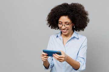 Young fun employee business corporate lawyer woman of African American ethnicity in classic shirt work in office use play racing app on mobile cell phone gadget smartphone isolated on grey background.