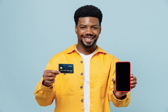 Young Man Of African American Ethnicity In Yellow Shirt Use Mobile Cell Phone Black Screen Workspace Area Credit Bank Card Shopping Online Order Delivery Isolated On Plain Pastel Light Blue Background