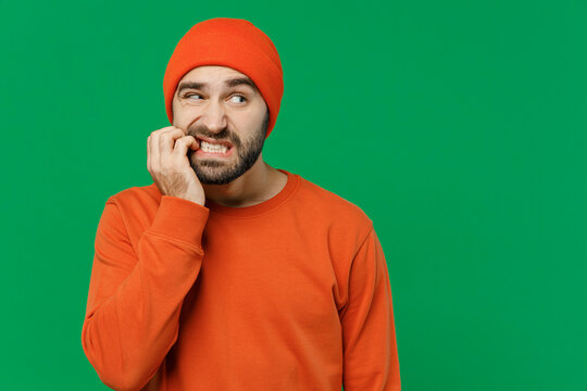 Young sad puzzled mistaken confused caucasian man 20s wear orange sweatshirt hat look aside biting fingers nails say oops isolated on plain green background studio portrait. People lifestyle concept.
